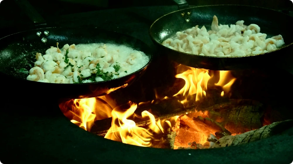 Two frying pans with food cooking over an open flame, showing a high-temperature kitchen environment.
