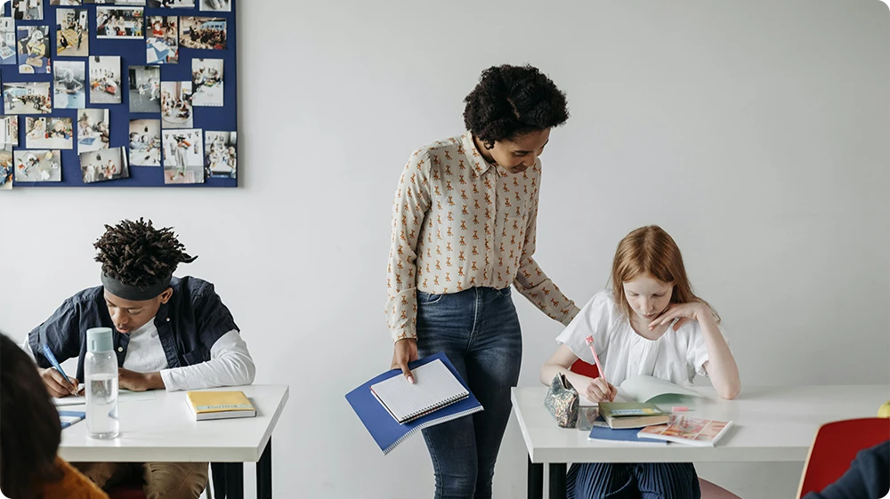 A teacher assisting a young student in a classroom while another student works independently; a photo board with pictures is visible in the background