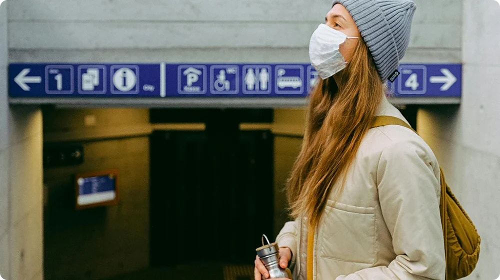  A young woman wearing a grey beanie and a white face mask stands near an underground station entrance, holding a reusable cup. She has long hair and a mustard yellow backpack. Above the entrance are directional signs indicating platforms, parking, restrooms, and other facilities.