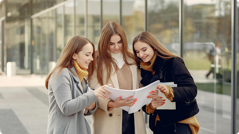 Three young women walking on a college campus carrying notebooks, representing students in further or higher education, such as A-levels, vocational courses, or degrees.