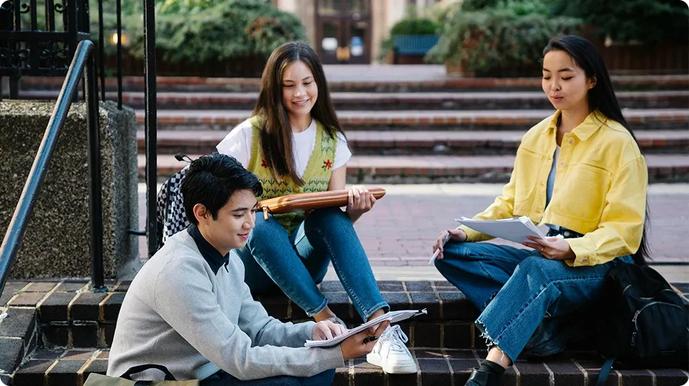 Group of diverse college students studying outdoors on campus steps, reading notes and smiling while sitting on brick stairs in a relaxed academic setting.