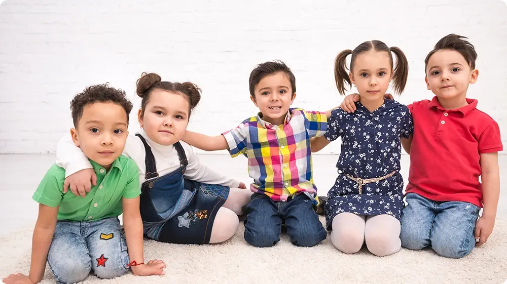 A group of seven diverse young children sitting in a row on the floor in front of a white brick wall, smiling and appearing joyful. They represent different ethnic backgrounds and are dressed in colourful casual clothes, reflecting inclusivity and unity in early childhood education.