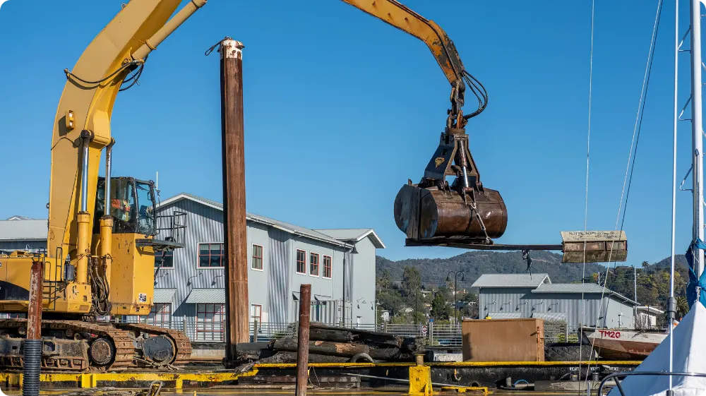 A large excavator with a grabber attachment on a dock, illustrating a heavy-duty lifting operation on a construction site.
