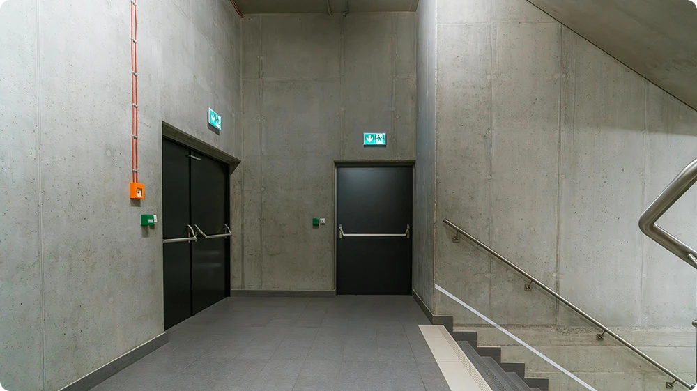 A modern emergency exit area in a commercial building, featuring two fire doors with illuminated green exit signs, a stairwell with a stainless-steel handrail, and wall-mounted emergency controls.
