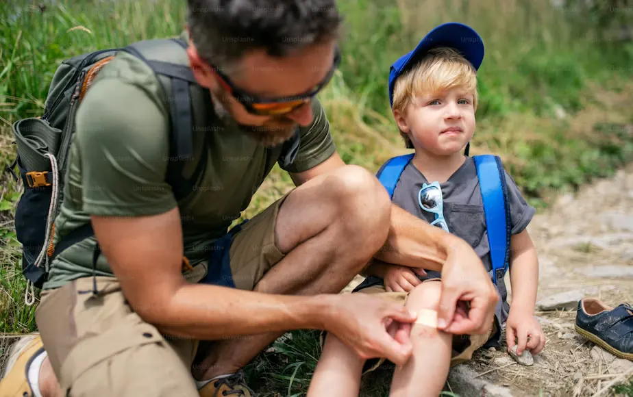 A father applying a plaster to his son's cut knee during an outdoor hike, demonstrating basic first aid for cuts and grazes.