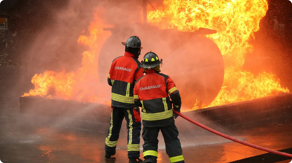 Firefighters in protective gear using hoses to extinguish a large blaze during fire safety training, representing UK workplace fire safety awareness