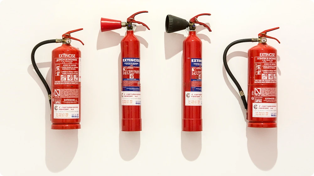Four red fire extinguishers mounted on a white wall, each with different nozzle types and usage labels, indicating variations such as CO and dry powder extinguishers.
