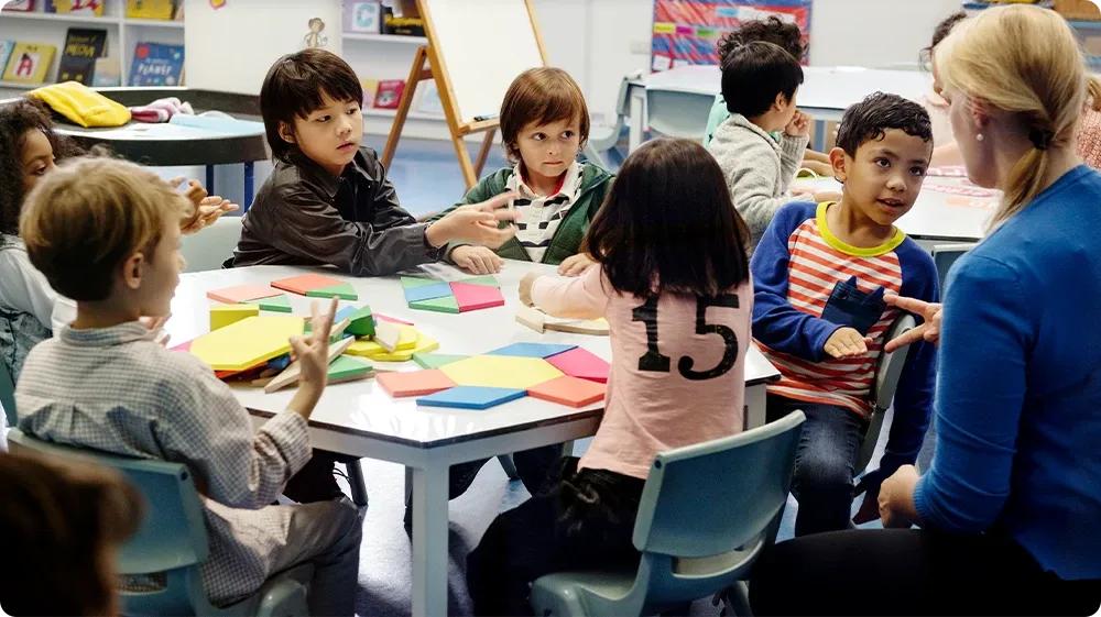 A diverse group of schoolchildren and their teacher gathered around a large table in a library or classroom, actively engaging with maps and books during a geography or world cultures lesson. The scene reflects inclusive education, collaborative learning, and multicultural understanding.