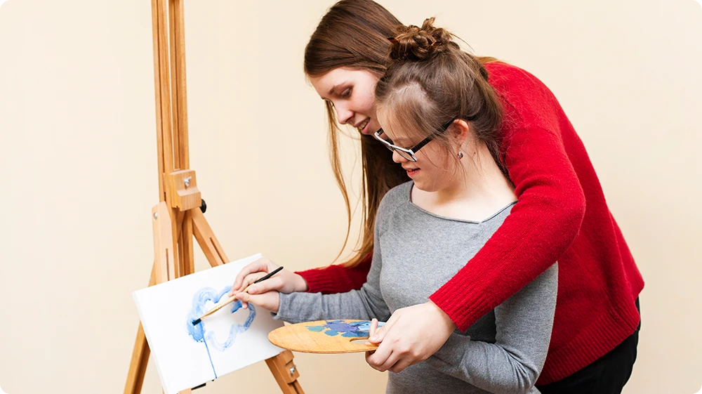  A teacher supports a young girl with Down syndrome as she writes on a chart pinned to a classroom chalkboard. The teacher gently guides her hand, demonstrating inclusive teaching practices and personalised learning support. 