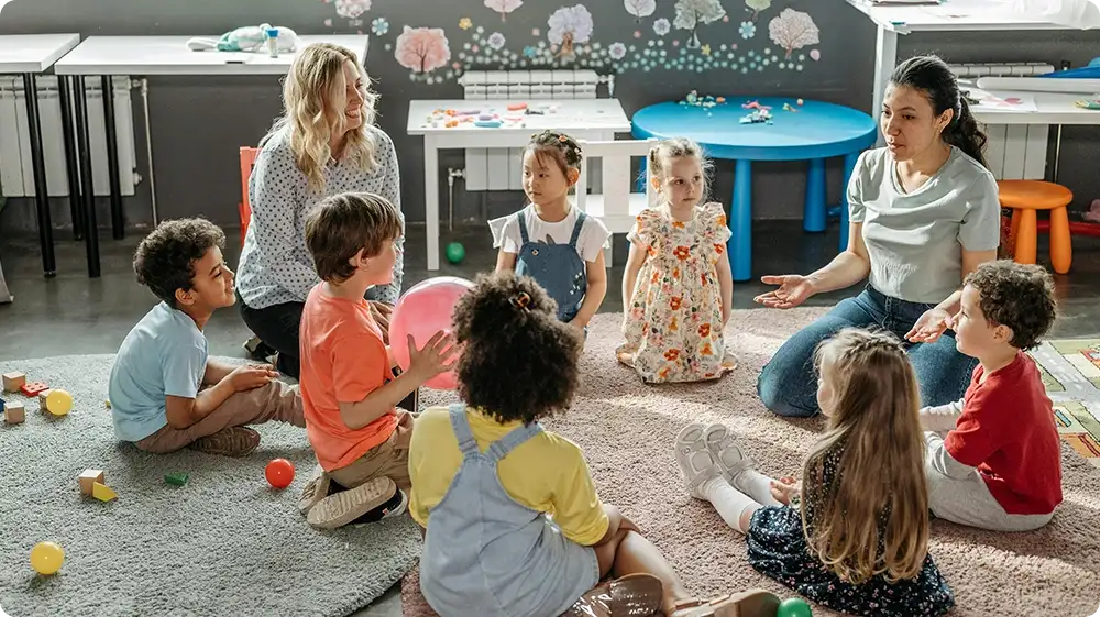 Two female teachers sitting in a circle with a group of young children in a colourful classroom, engaging in a group activity or discussion on a rug surrounded by toys.