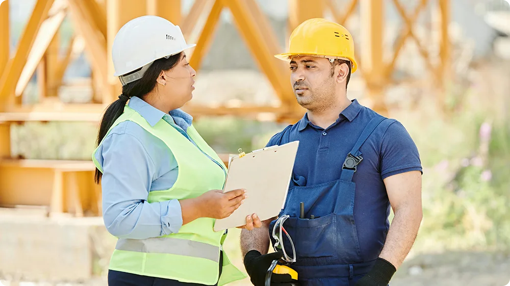 Two construction site managers discussing safety procedures on-site, wearing PPE and hard hats during site safety training.