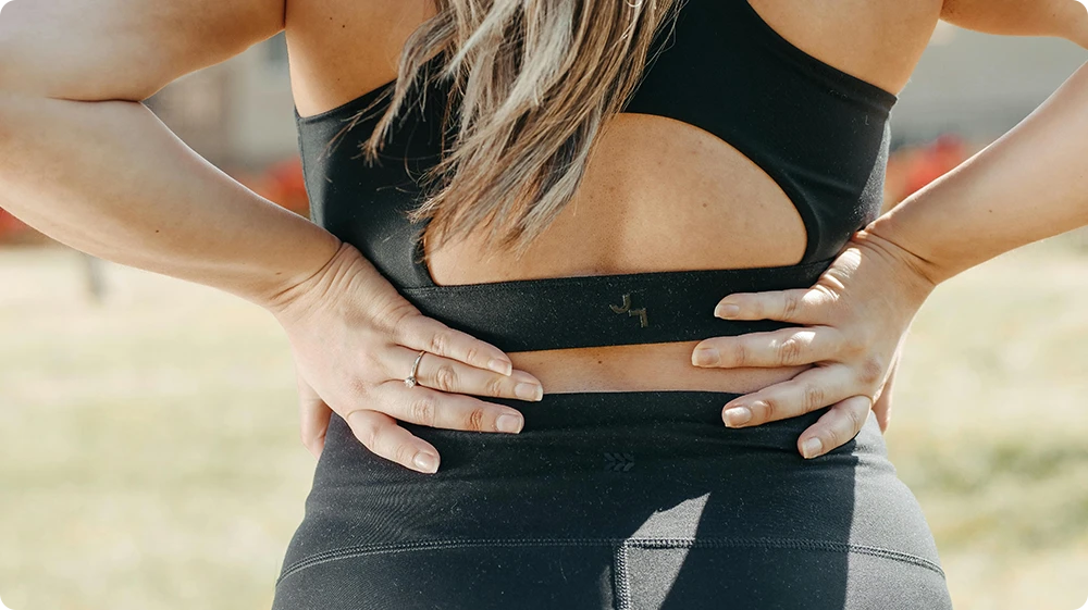 A woman standing with her hands on her lower back, showing signs of spinal discomfort or injury