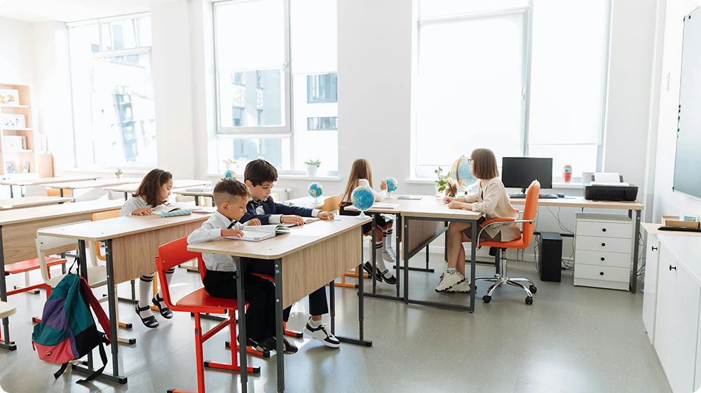 Teaching assistant observing primary school children working independently at their desks in a bright, modern classroom environment.