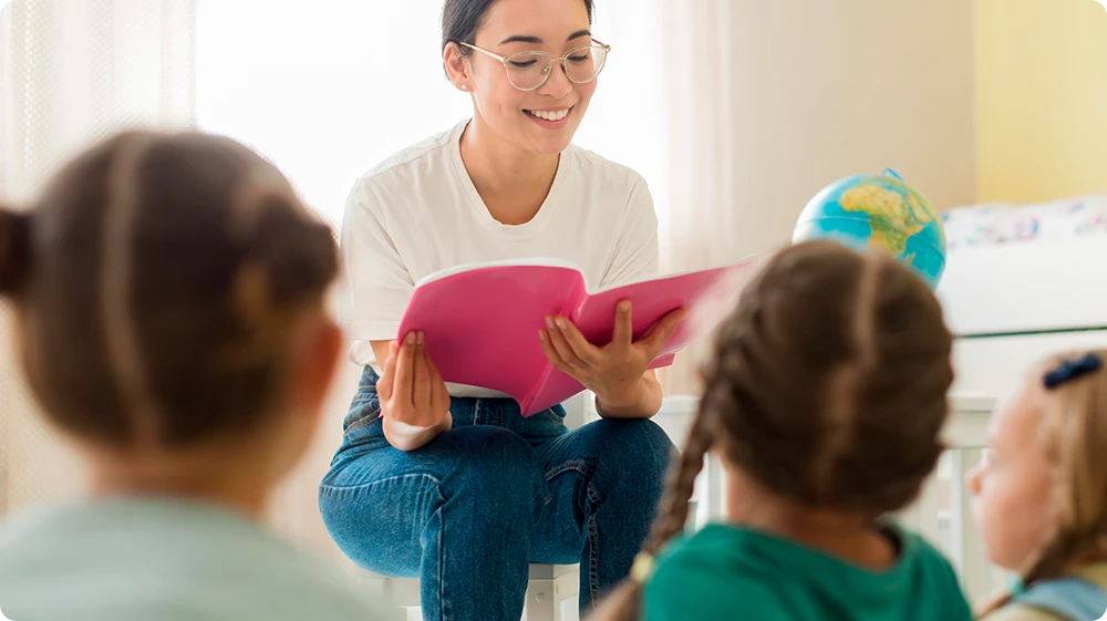 A teaching Assistant sits and engages with a group of young children in a classroom, holding a clipboard while they listen attentively, with a chalkboard and learning materials in the background.