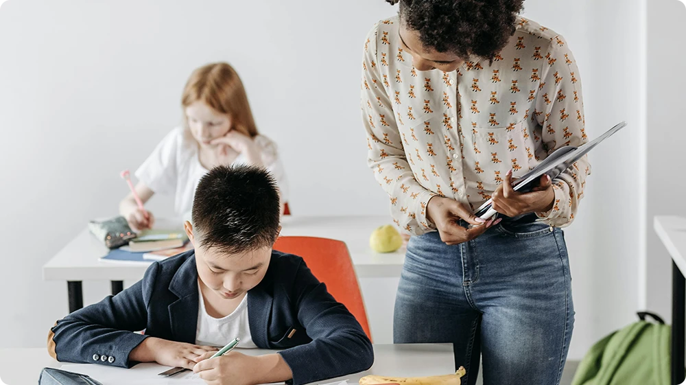 Female teacher or teaching assistant observing students working at their desks in a bright, modern classroom setting.