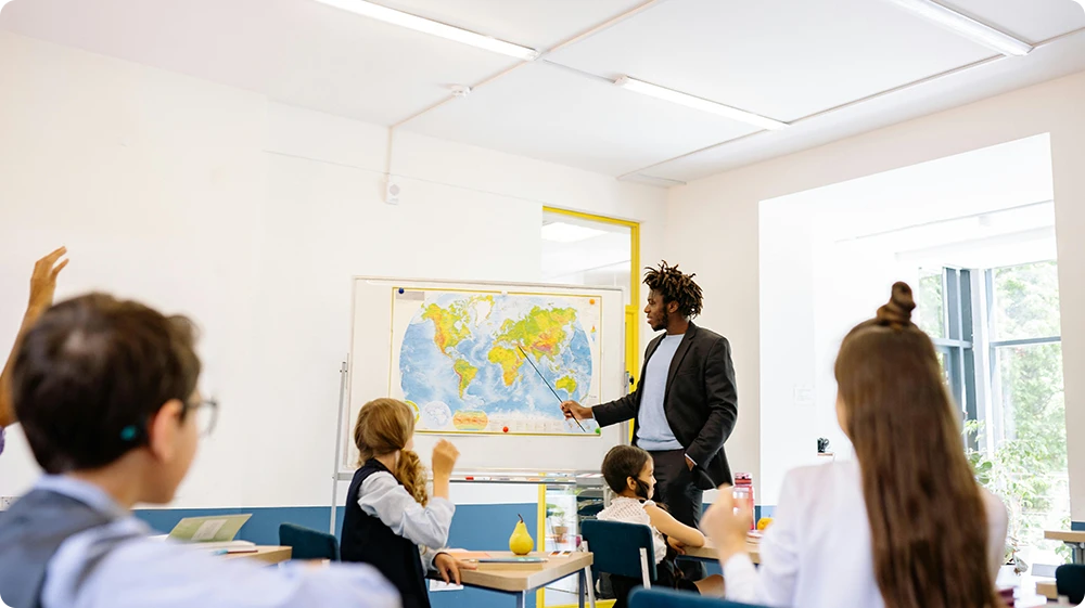A teacher pointing at a world map during a geography lesson while students seated at desks attentively listen and participate in a classroom setting