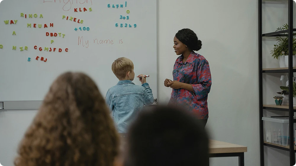 Teaching assistant helping a primary school student write on the whiteboard during an English lesson, with other children observing in a classroom setting.