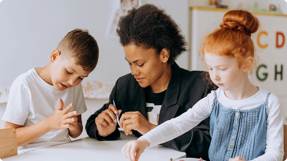 Teaching assistant engaging with young children during a hands-on learning activity in an early years classroom, showcasing creative play, early childhood education, and inclusive learning support.