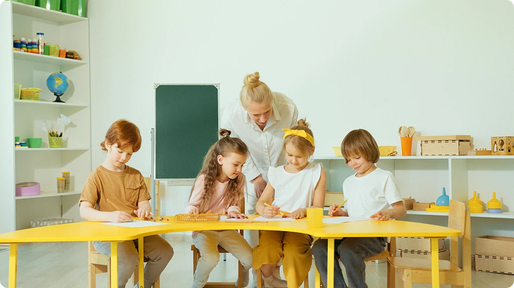 Teaching assistant supporting a group of primary school children with a classroom activity at a yellow table in an early years learning environment.