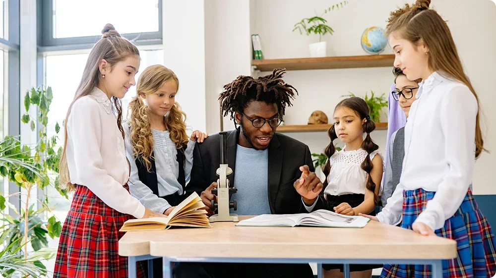 A teaching assistant explaining science concepts using a microscope to a group of primary school girls in uniform during a hands-on classroom activity.