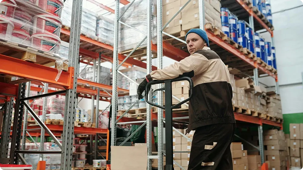 A warehouse worker in a brown uniform and blue beanie standing behind a manual pallet jack, surrounded by tall shelves stacked with boxes, paint buckets, and industrial supplies.