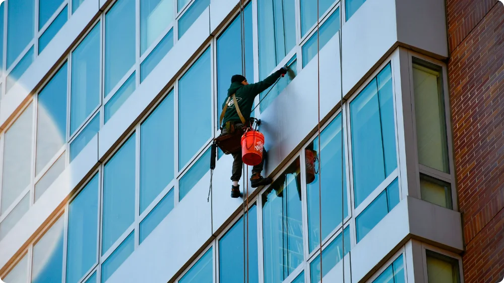 A window washer, securely attached to safety ropes, cleans the exterior glass of a modern multi-story building