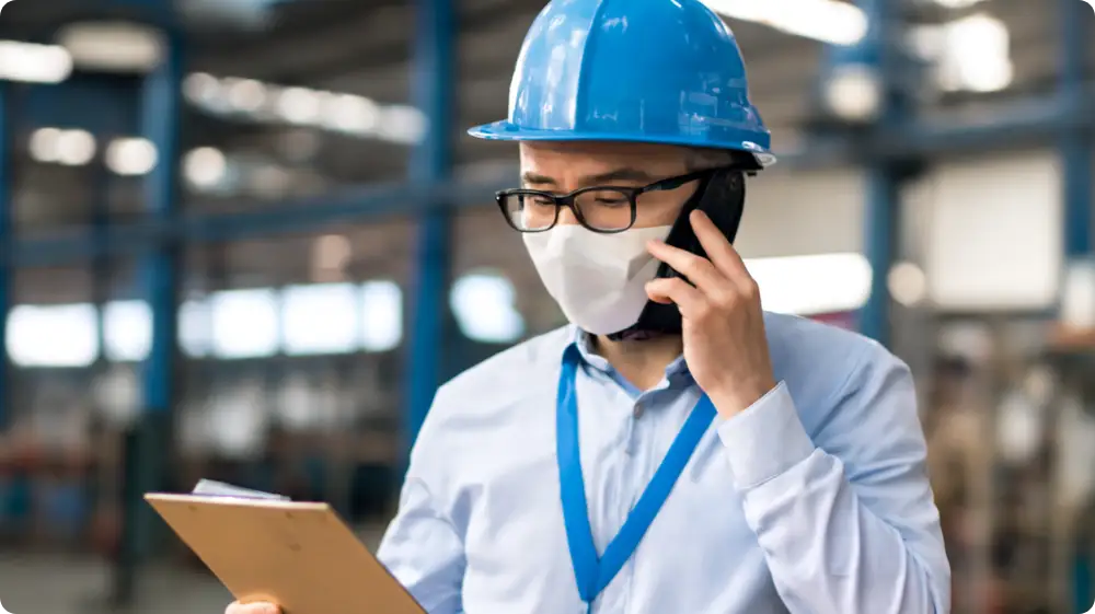 An Engineer wearing a hard hat and a face mask, holding a file in a workplace environment.