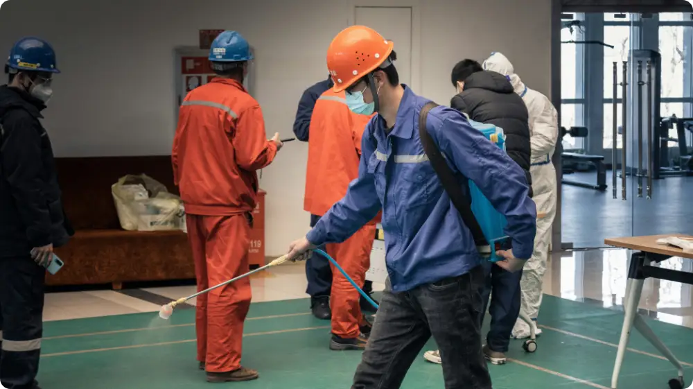 A worker in a blue uniform is spraying disinfectant in a workplace environment, wearing a helmet and mask as part of safety protocols.