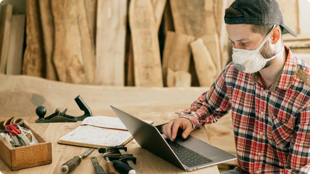 Carpenter using a laptop in a wood workshop for the NVQ Level 2 Carpentry assessment and certification process