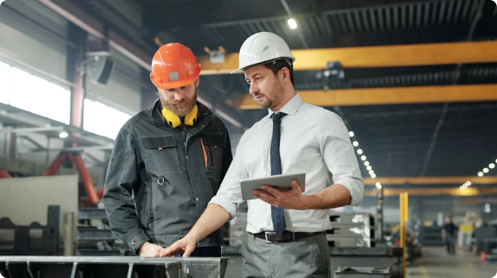 A construction manager and a site worker wearing safety helmets review project details on a tablet inside a manufacturing or industrial facility.