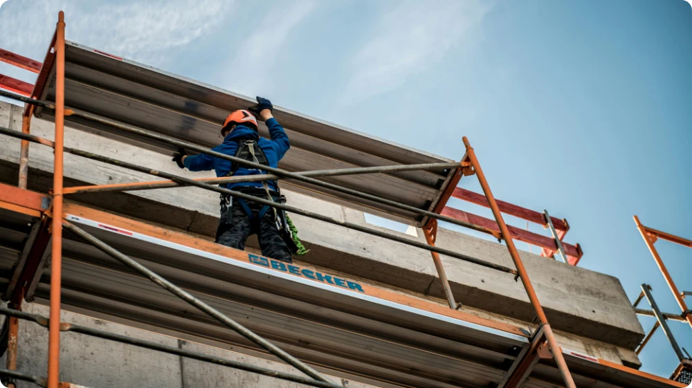 A construction worker in a hard hat and safety harness stands on a large scaffold, working on the exterior of the building.