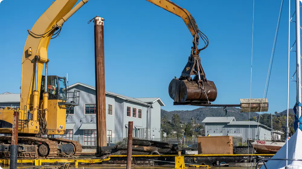 A large excavator with a grabber attachment on a dock, illustrating a heavy-duty lifting operation on a construction site