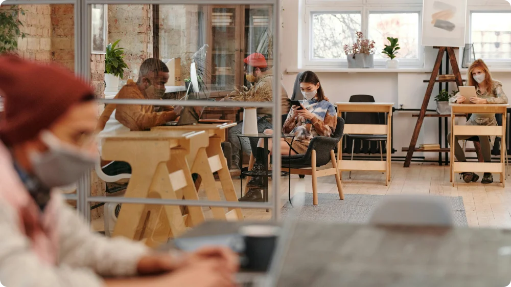 People wearing face masks work and sit apart in a modern, socially distanced office space with glass partitions and wooden desks.