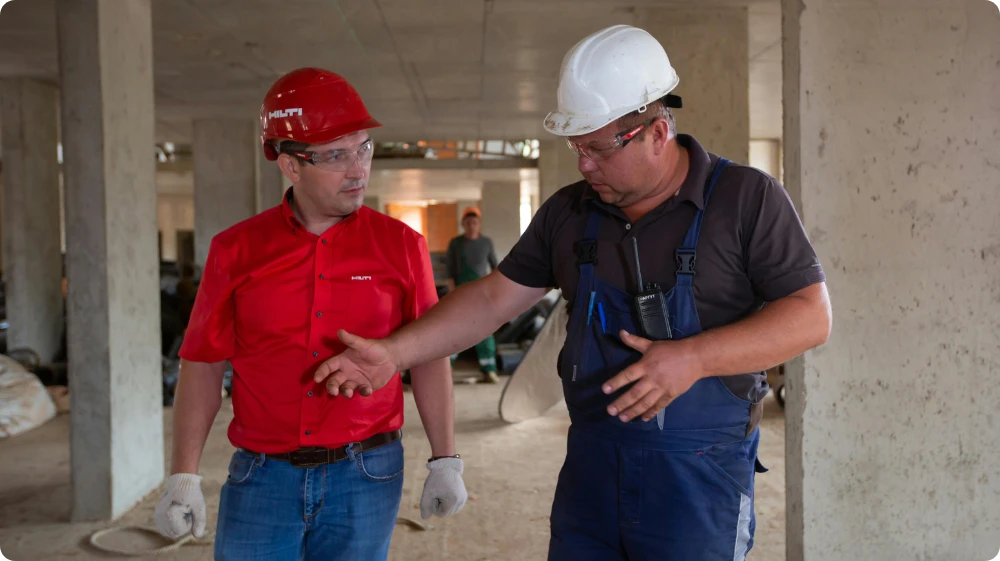 Two male workers wearing safety helmets inspect components on a factory floor