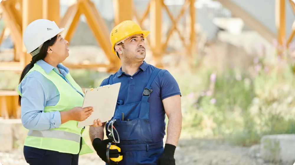 A male and female worker wearing hard hats and safety vests, inspecting a clipboard near a large crane, representing a safety inspection. 