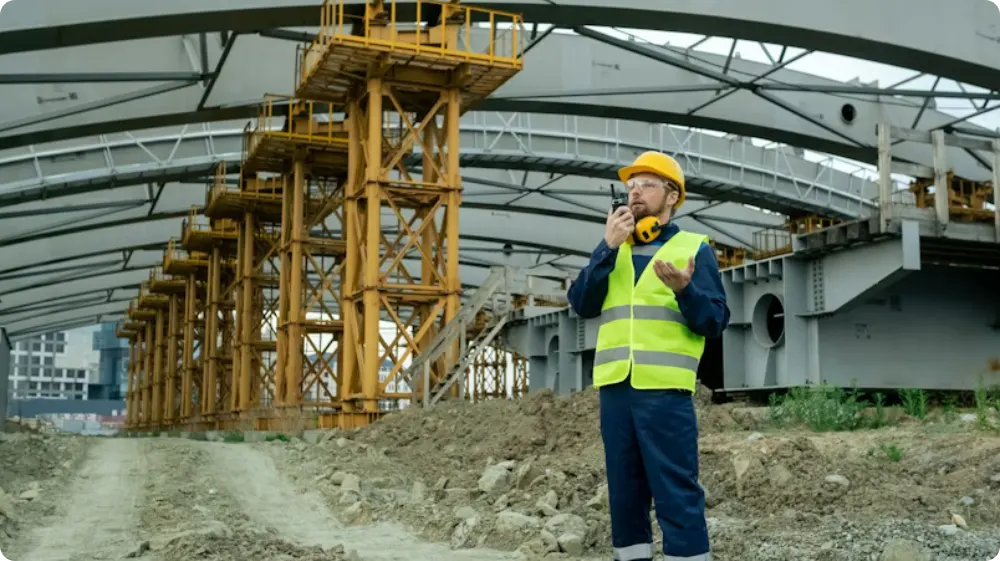 Construction site supervisor wearing a yellow hard hat and high-visibility vest, using a walkie-talkie while overseeing structural steelwork and scaffolding at an industrial site.