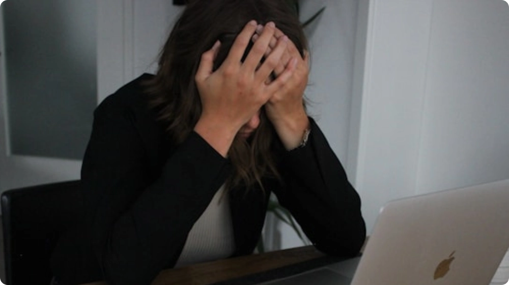 Stressed woman sitting at a desk with her head in her hands while working on a laptop, representing workplace stress