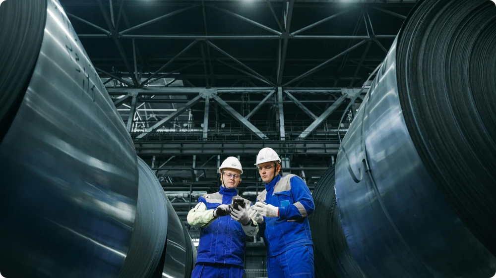 Two employees at a factory, wearing helmets and vests, showcase their use of personal protective equipment (PPE) at their workplace