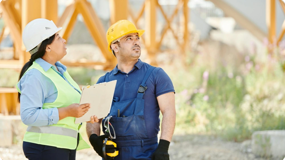 A male and female worker wearing hard hats and safety vests, inspecting a clipboard near a large crane, representing a safety inspection.