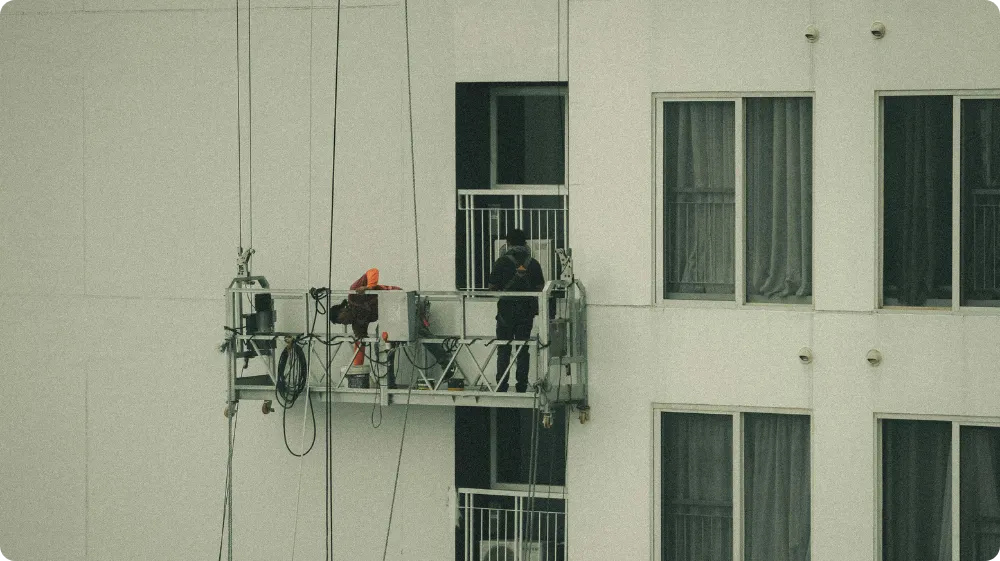 Two workers in safety harnesses and helmets are lowered from the top of the building to work on the balcony